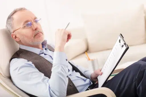 Older man in glasses thinking while reviewing documents on a clipboard during a planning discussion.