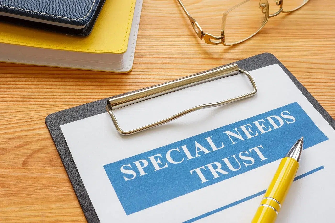 A clipboard labeled “Special Needs Trust” on a wooden desk with a yellow pen, notebooks, and eyeglasses.