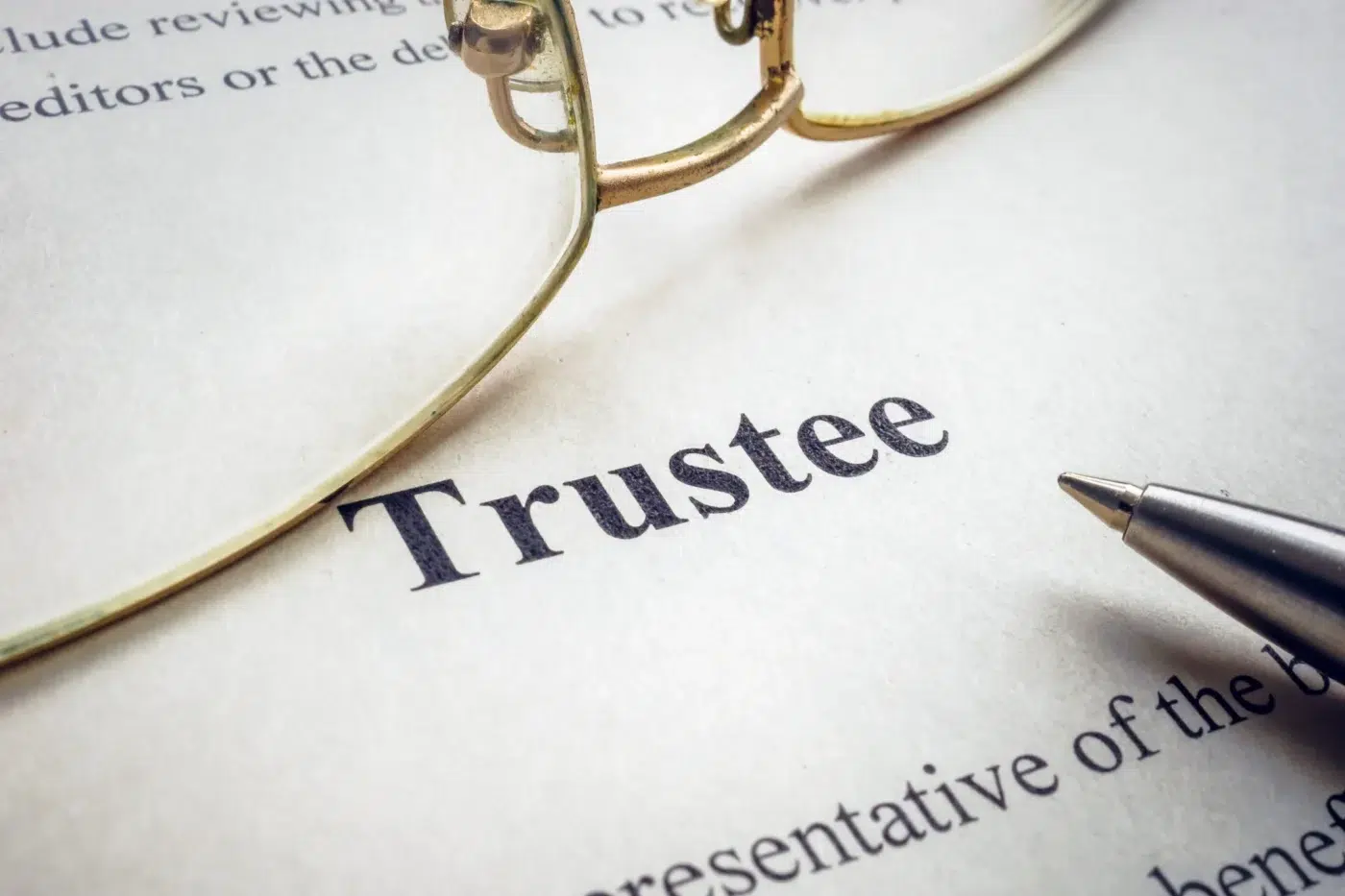 A close-up of the word “Trustee” printed on legal paperwork with eyeglasses and a pen resting nearby.