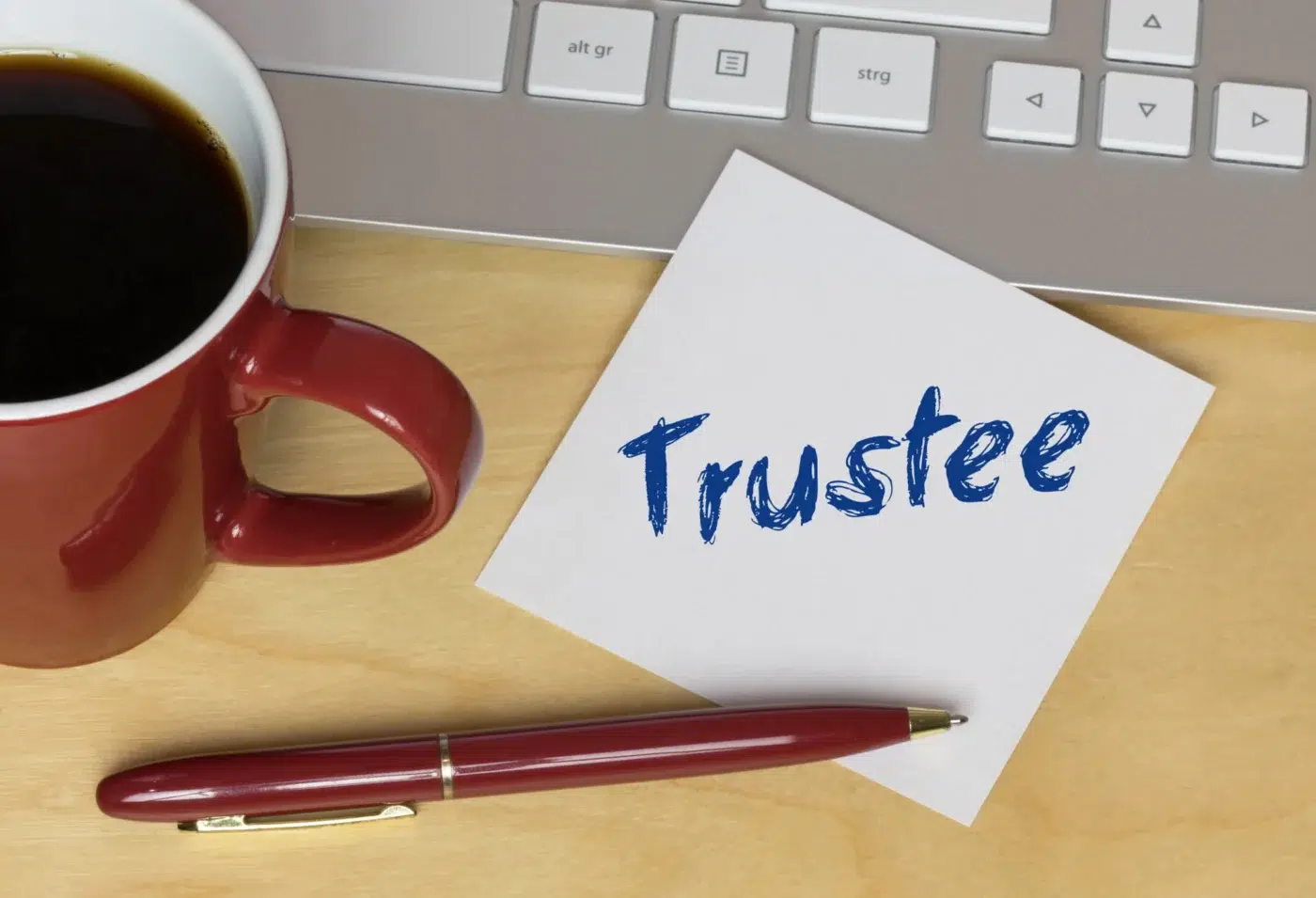 A desk with a red coffee mug, a red pen, a keyboard, and a note card labeled “Trustee.”